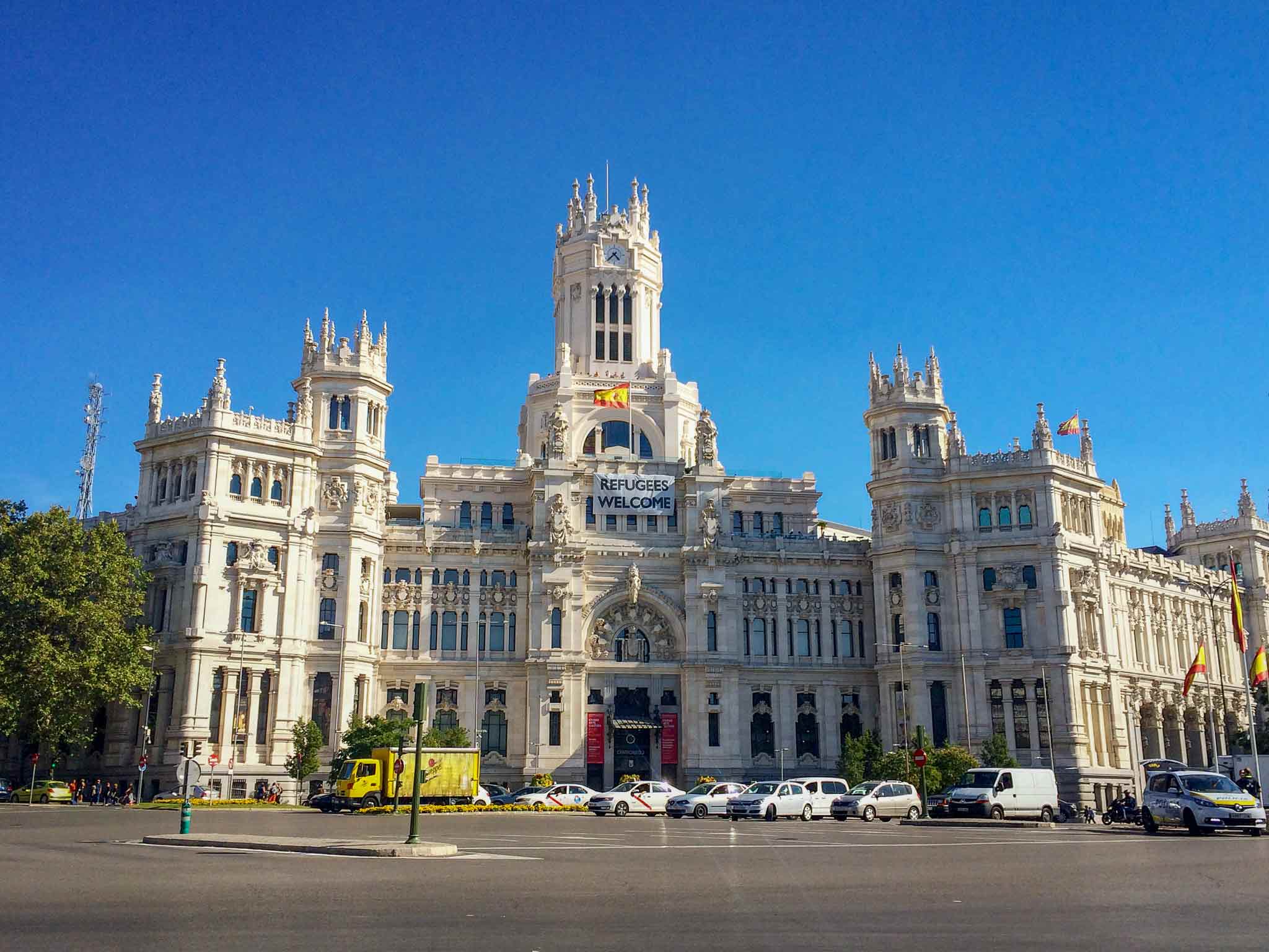 Picture of Cibeles Palace with a Spanish flag blowing in the wind and a refugees welcome sign in Madrid, Spain.