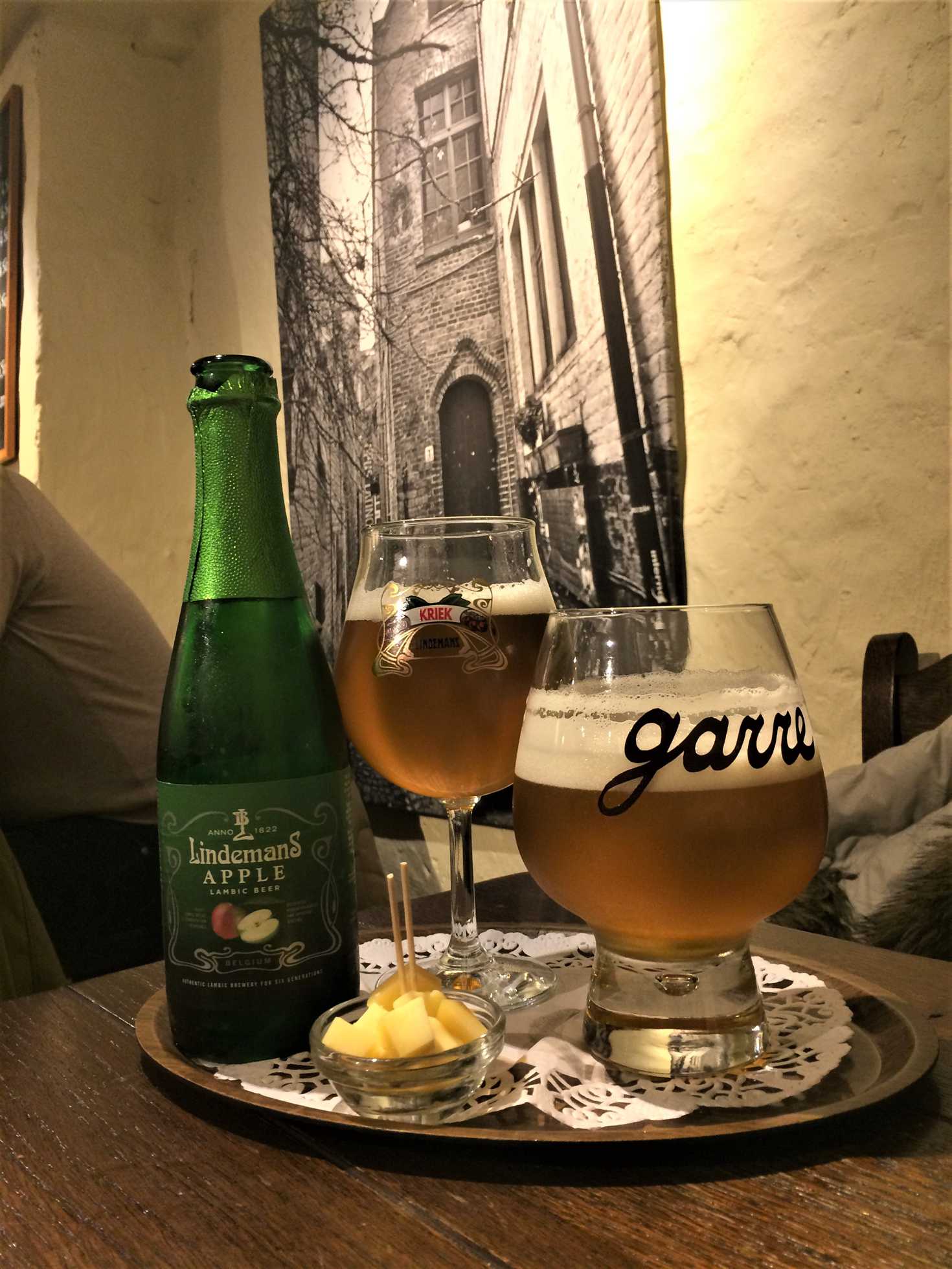 A picture of two glasses of beer, a bottle of lindman's apple lambic beer, and a small dish filled with cubed cheese.