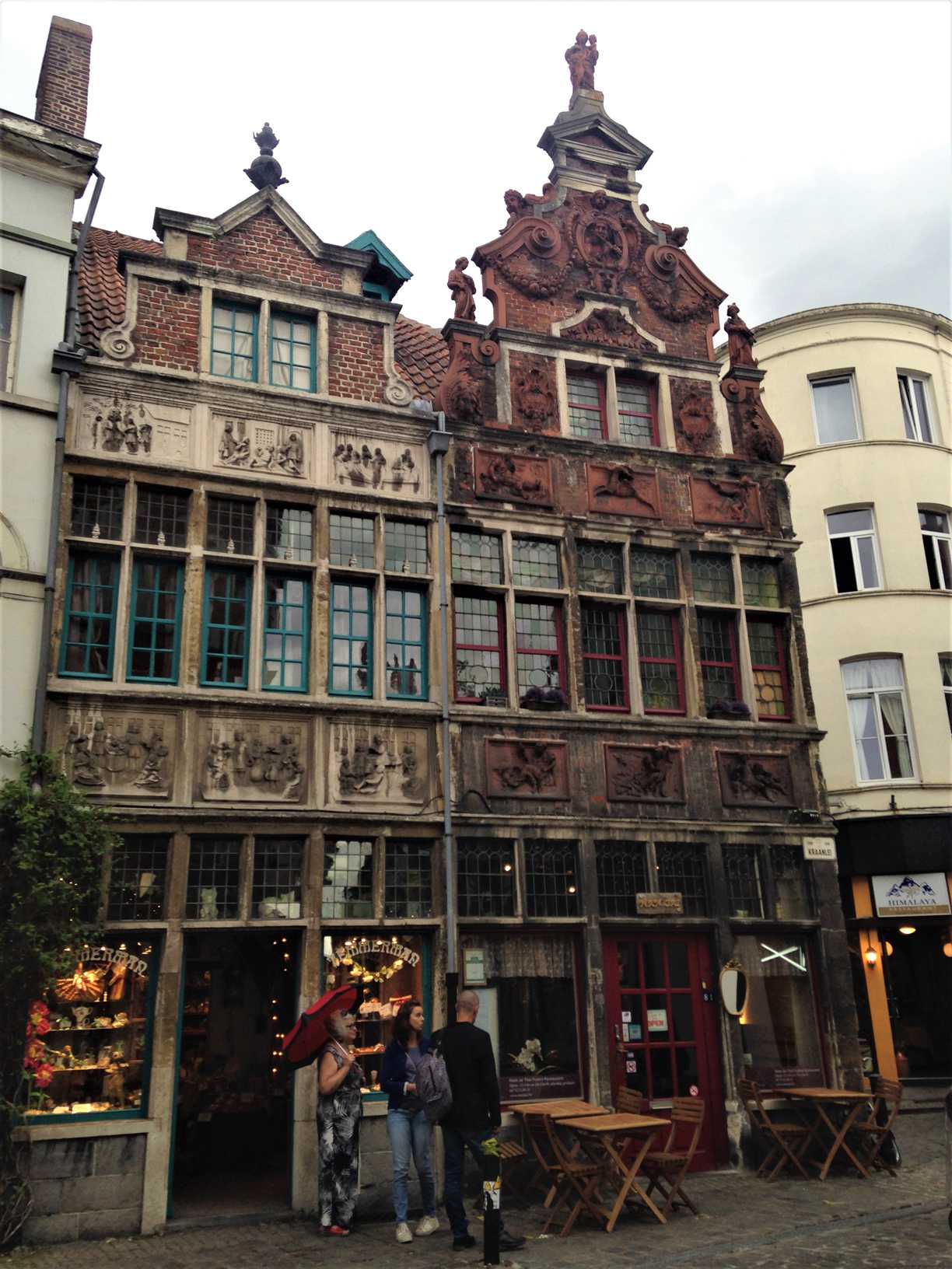 A picture of a unique brick building in Ghent, Belgium with stone carvings on the facade and gables. Tables and chairs are outside of the building, which is on a cobblestone street.