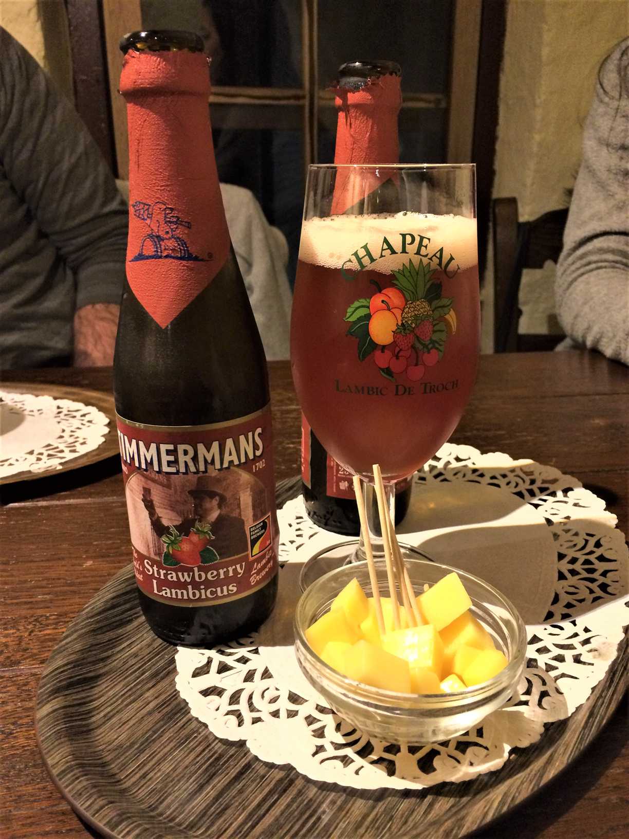This picture shows two bottles of Zimmermans strawberry lambicus beer, a glass of beer, and a small bowl of cubed cheese with toothpicks sticking out.