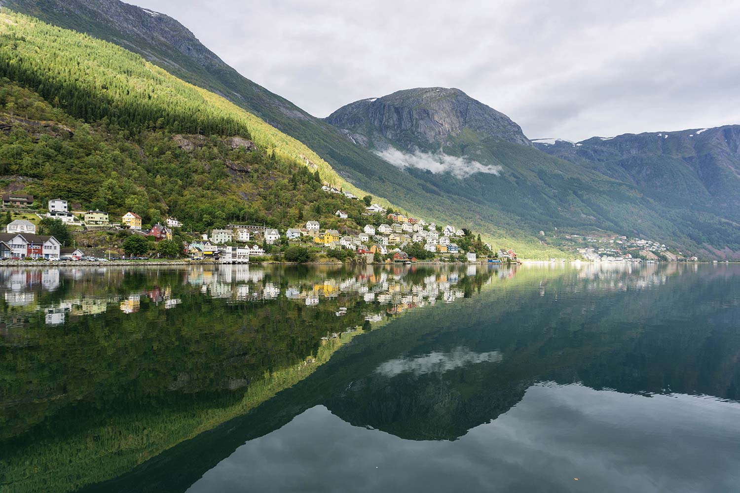 A picture of the town of Odda, Norway and its reflection with mountains and clouds in the background.