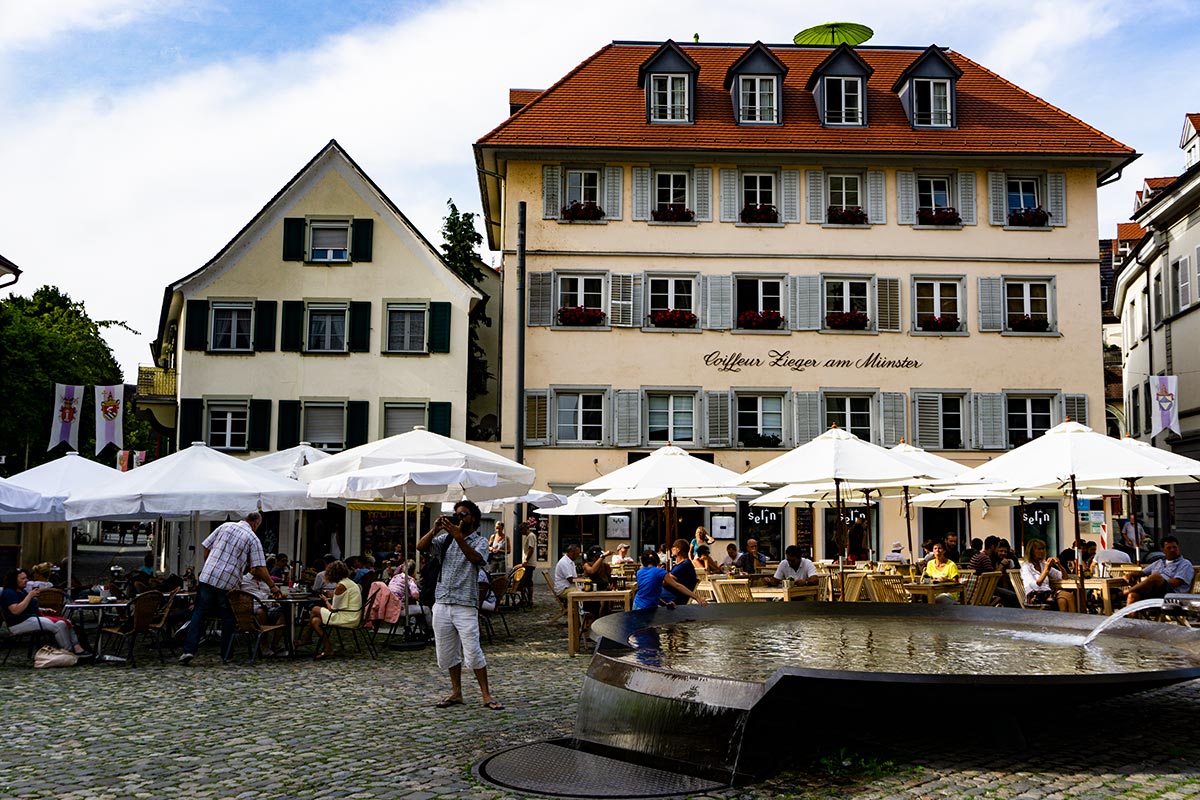 A plaza in Meersburg, Germany with a fountain and tables with umbrellas.