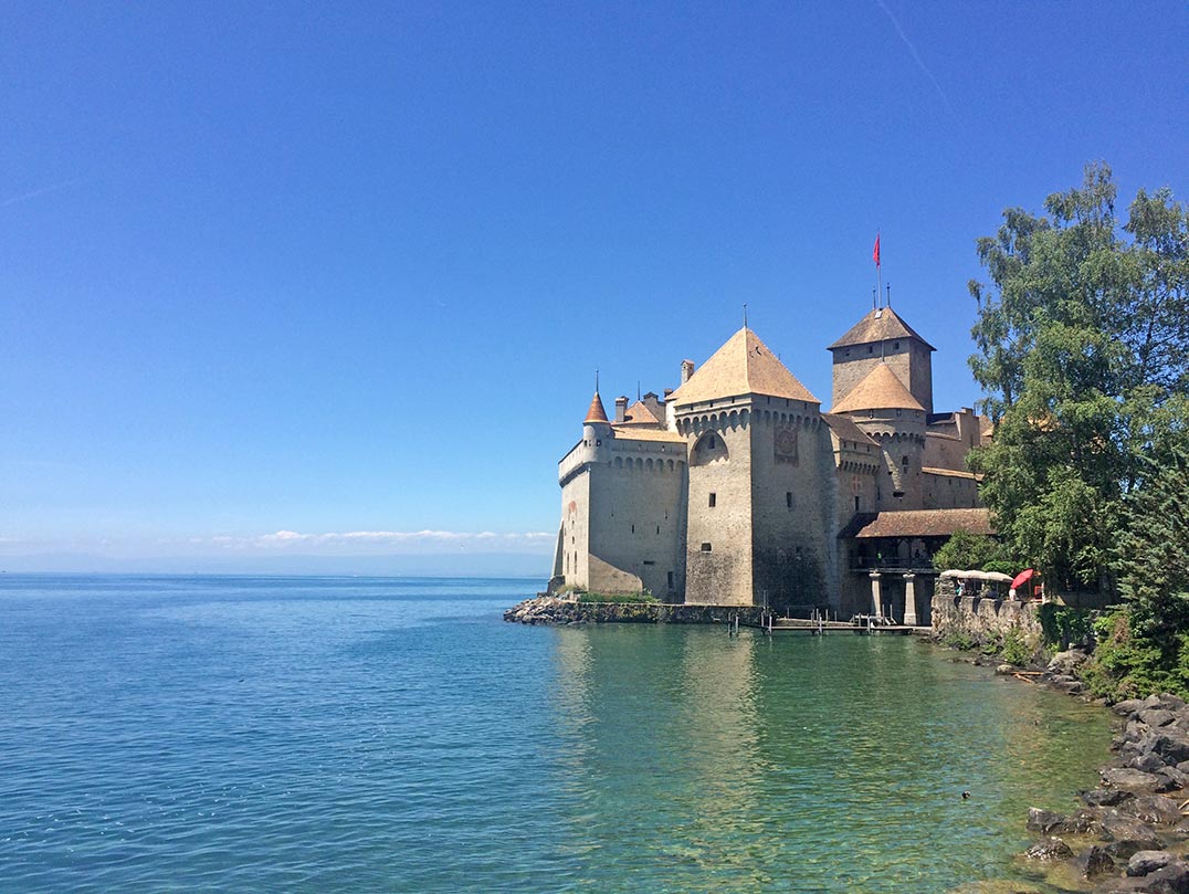 Chateau de Chillon in Montreux, Switzerland. The buidling is made of stone with a red roof and is situated right on the banks of Lake Geneva. The sky is a clear blue.